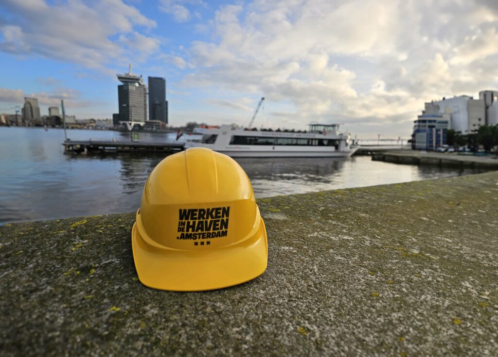 op de voorgrond ligt een gele veiligheidshelm met de tekst "werken in de haven.amsterdam"
in de achtergrond zie je de boot river dream